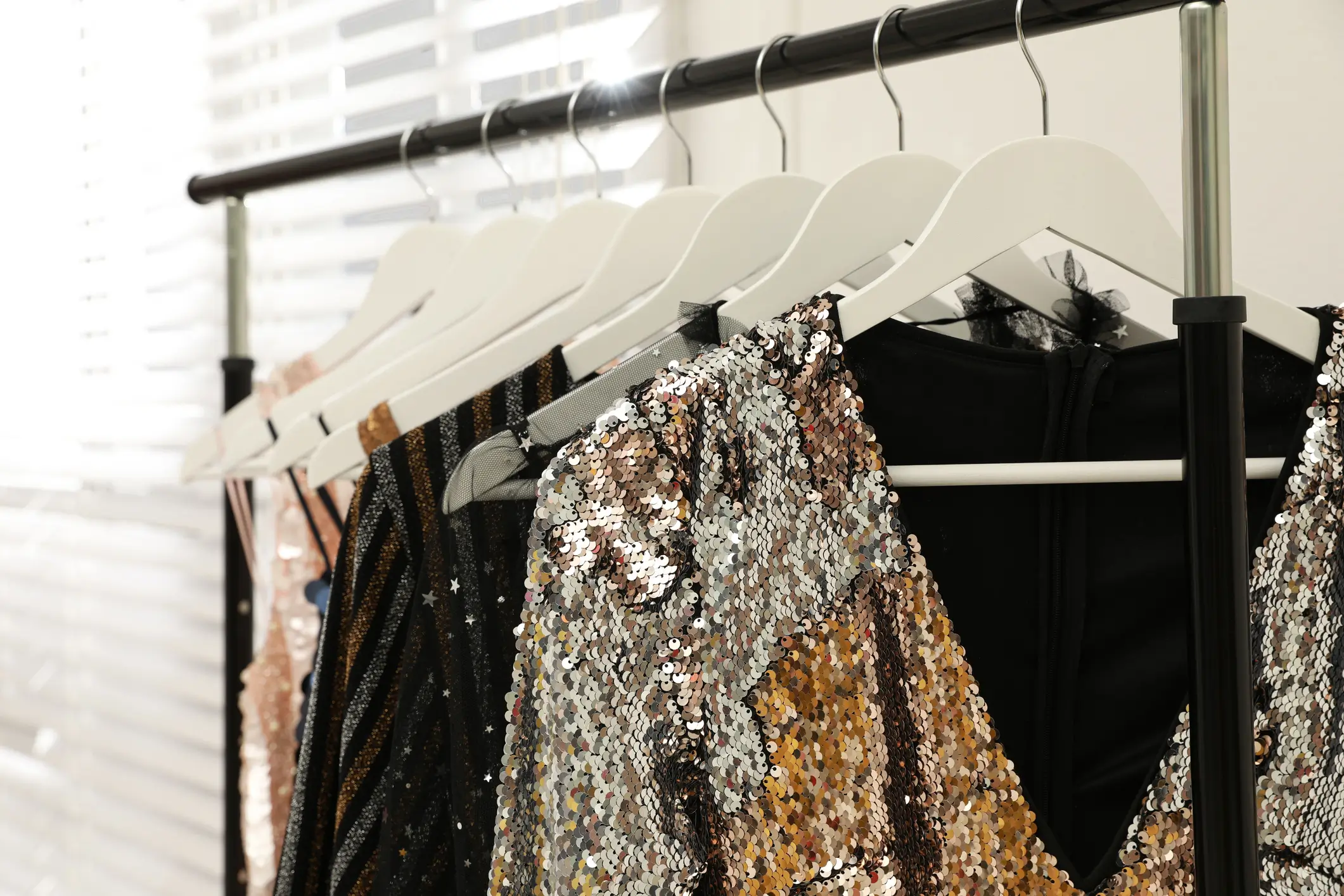 Five sequined dresses in gold, silver, and black hang on white hangers from a black clothing rack in a well-lit room with blinds.